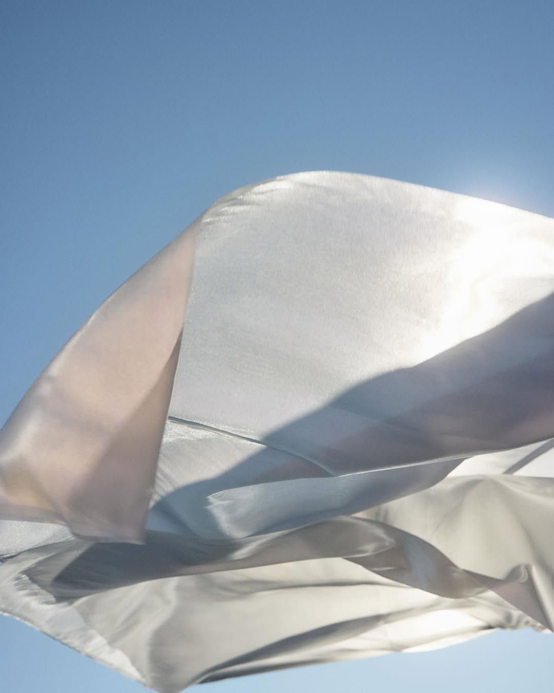 A light-colored fabric is billowing in the wind against a gradient blue sky with a visible sun glare, creating a calming effect.