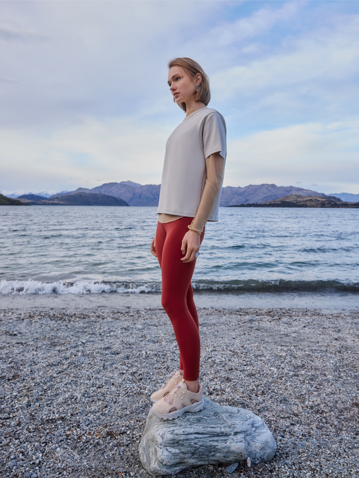 A woman in athletic apparel stands on a rock near a lake with distant mountains under a cloudy sky, captured in a medium, full shot.