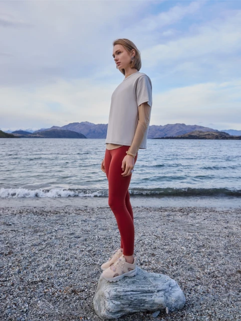 A woman in athletic apparel stands on a rock near a lake with distant mountains under a cloudy sky, captured in a medium, full shot.