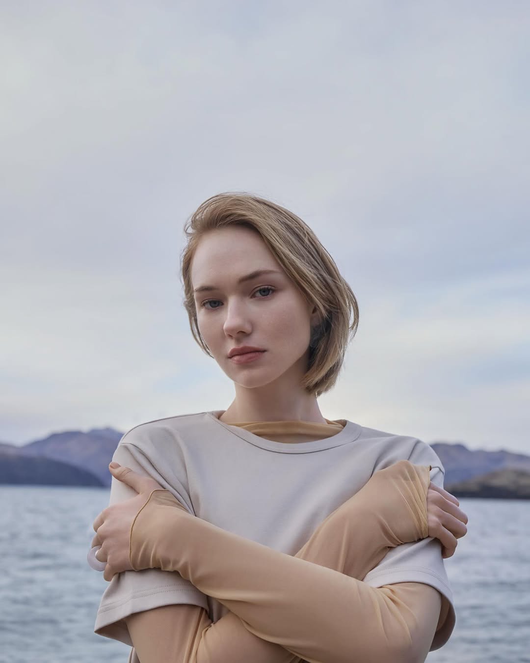 Portrait of a young woman with short blonde hair standing near a lake with distant mountains under an overcast sky, wearing casual clothing.