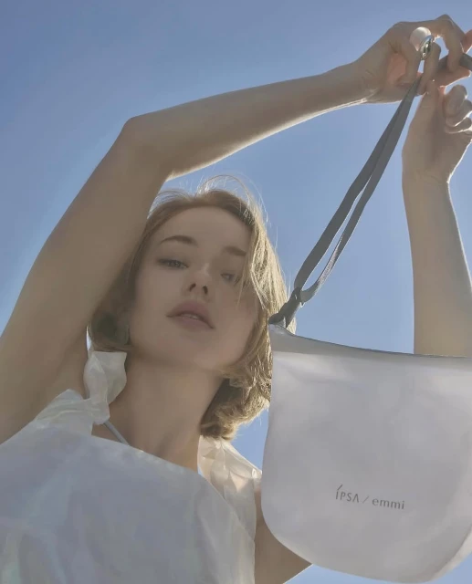 Woman holding a white IPSA/EMMI handbag, shot from a low angle against a bright blue sky in an outdoor setting with natural light.