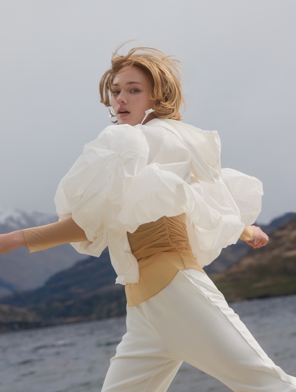 Young woman in a white blouse and pants poses outdoors, with a lake and mountains in the background. Fashion editorial style.