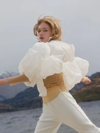 Young woman in a white blouse and pants poses outdoors, with a lake and mountains in the background. Fashion editorial style.