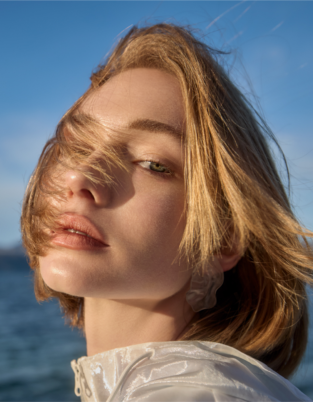 Close-up portrait of a fair-skinned woman with blonde hair, light make-up, and a translucent top against a blue sky and water.