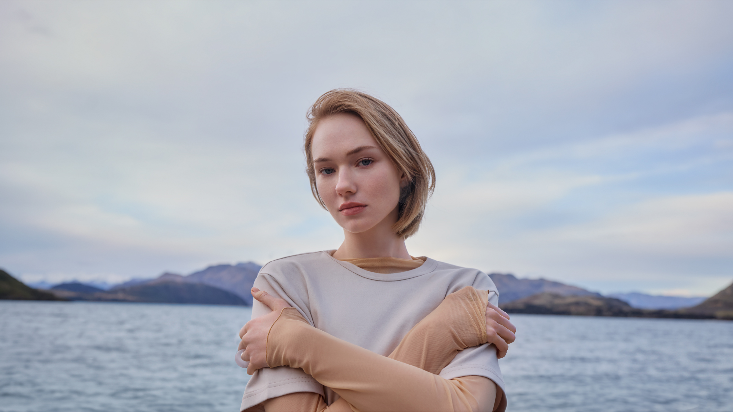 Young woman in a beige t-shirt poses outdoors with arms crossed at the lake edge, mountains and cloudy sky in the background.