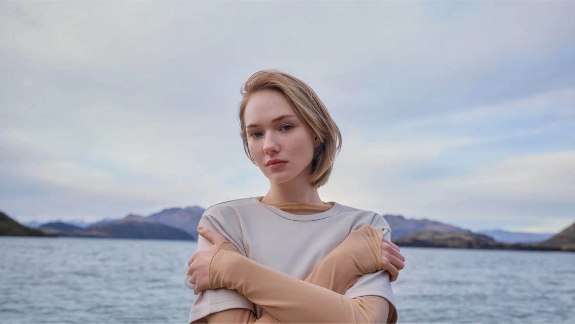Young woman in a beige t-shirt poses outdoors with arms crossed at the lake edge, mountains and cloudy sky in the background.