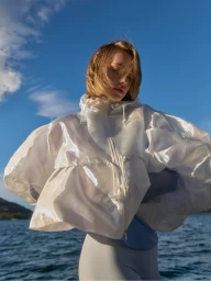 Model wearing a sheer white puffer jacket outdoors against a blue water and sky background by Ben Clement.
