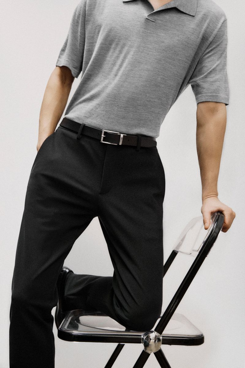 Man wearing a gray polo shirt and black pants poses while leaning on a clear plastic chair in a clean, neutral studio setting.