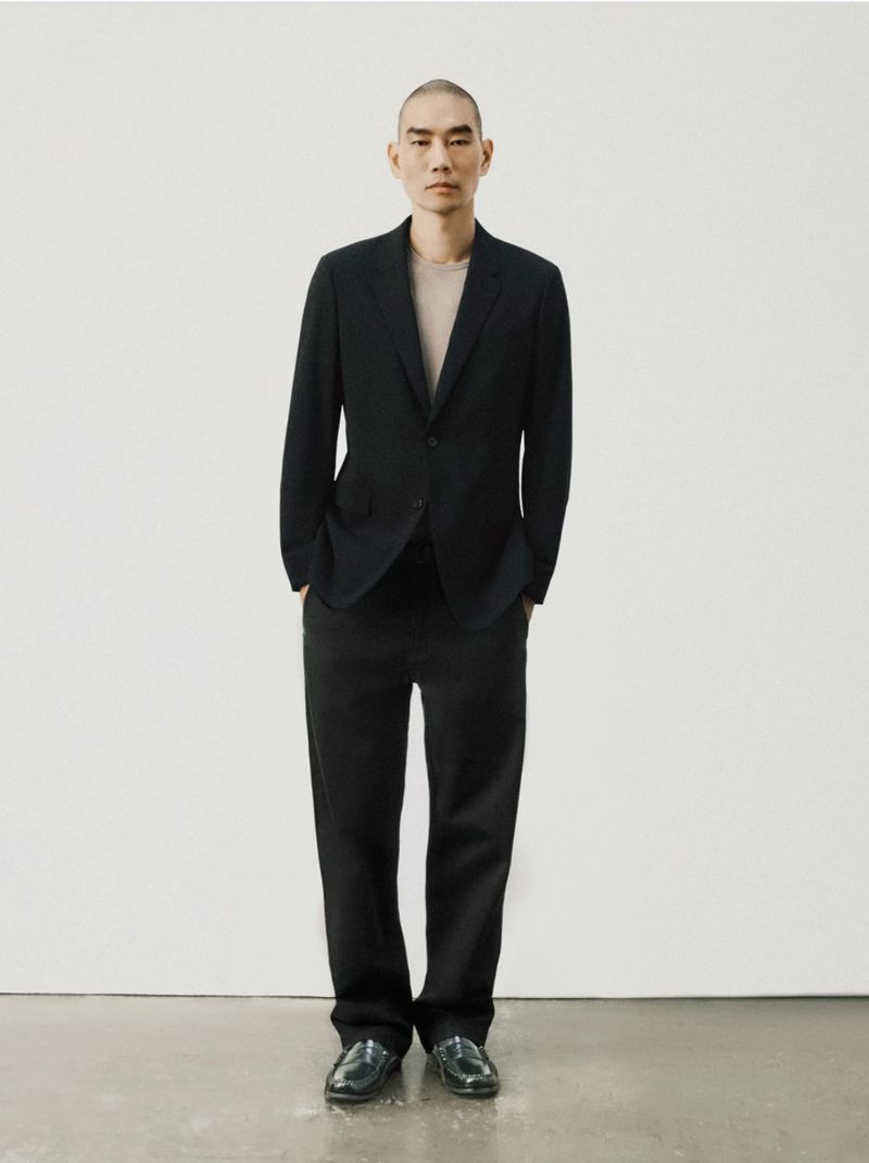 A full-length portrait captures an adult male in a dark suit against a white backdrop, showcasing a modern and minimalist aesthetic.