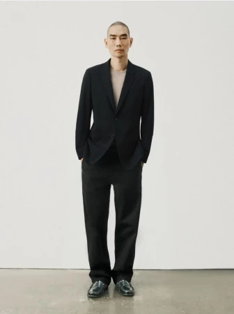 A full-length portrait captures an adult male in a dark suit against a white backdrop, showcasing a modern and minimalist aesthetic.