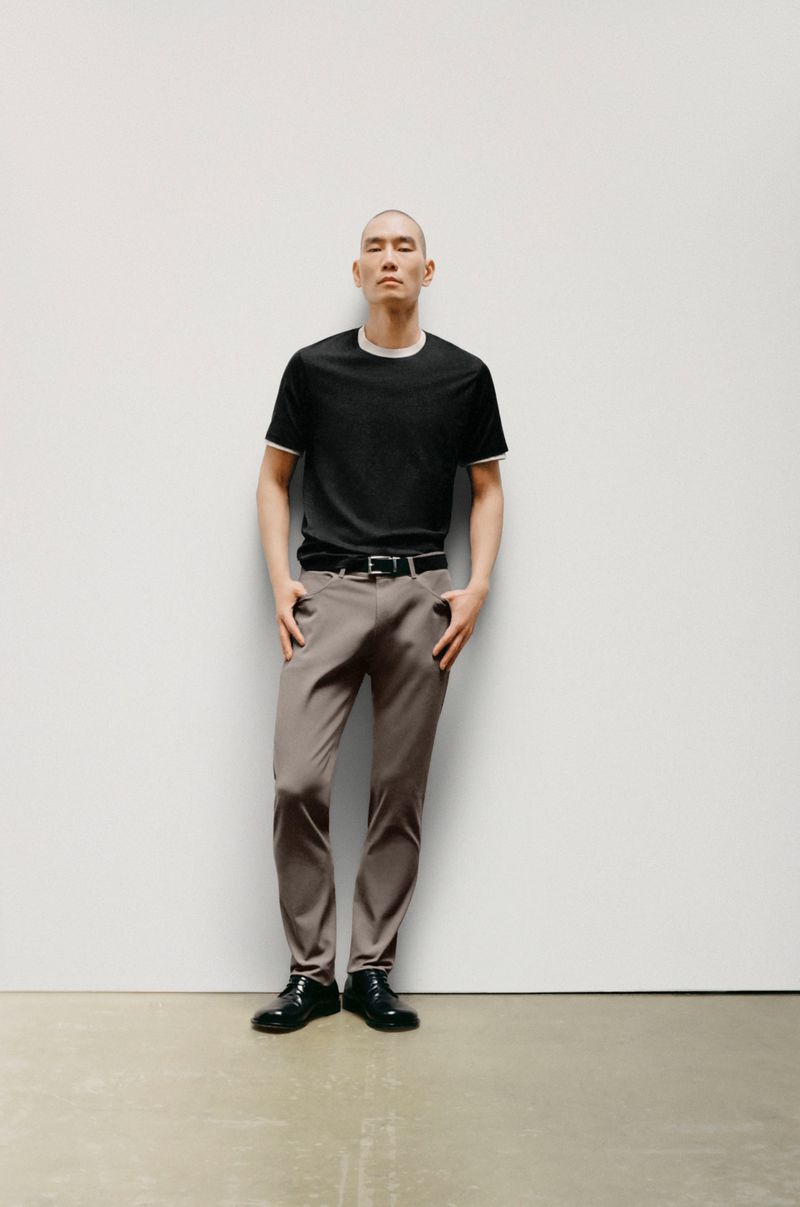 Full-length shot of a bald adult male posing in a studio, wearing a black t-shirt, gray trousers, a black belt, and black leather shoes.
