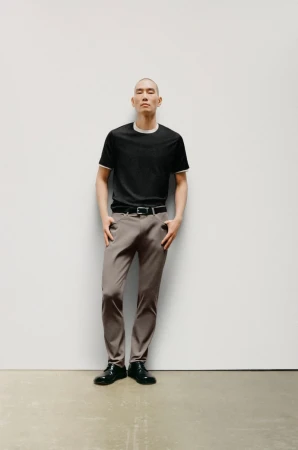 Full-length shot of a bald adult male posing in a studio, wearing a black t-shirt, gray trousers, a black belt, and black leather shoes.