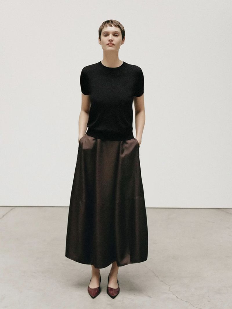 A full-length portrait of an adult woman in a black short-sleeved shirt and dark brown skirt, posing in a brightly lit neutral interior.