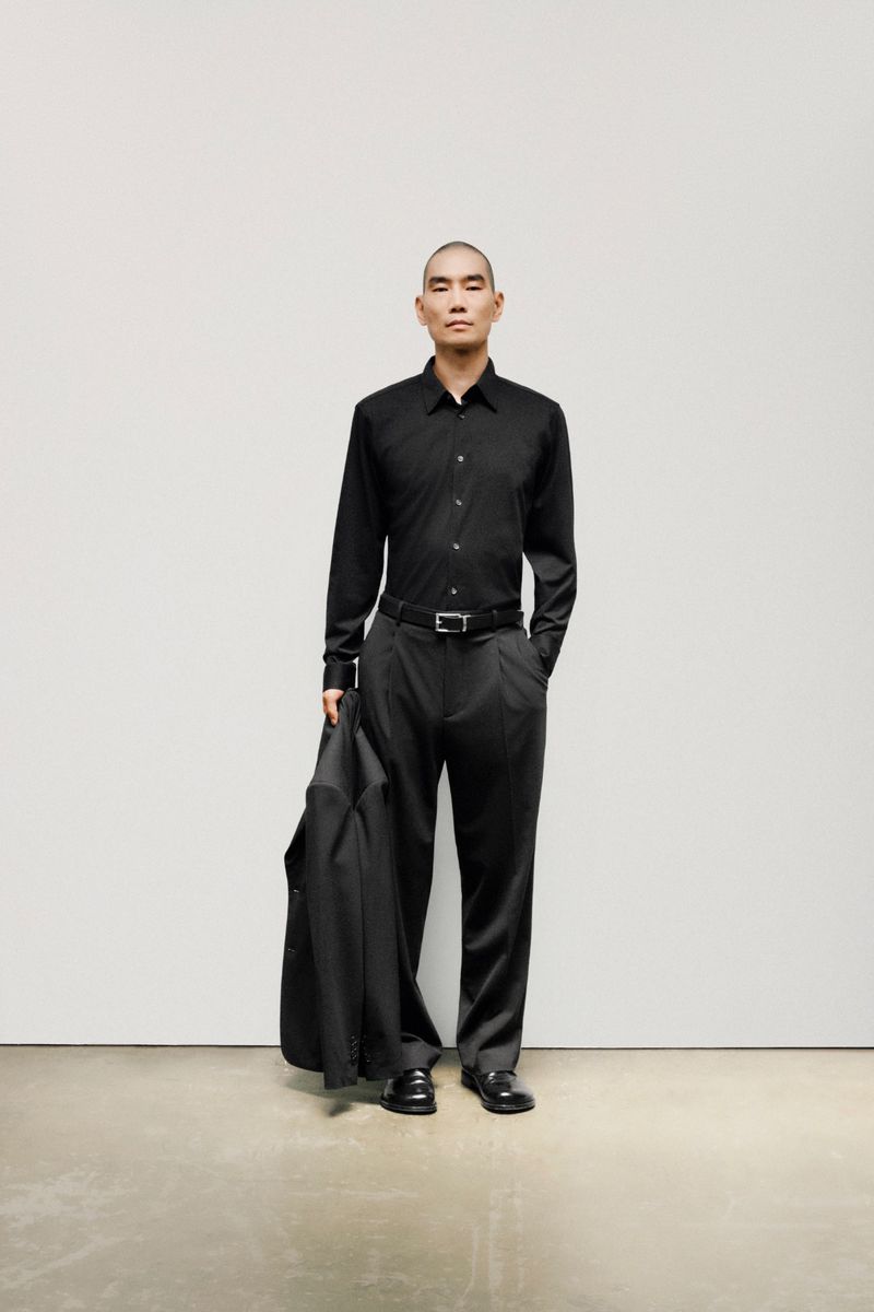 An adult Asian man with shaved head poses in an all-black satin button-down shirt, dress pants, loafers and holds a suit jacket in studio.