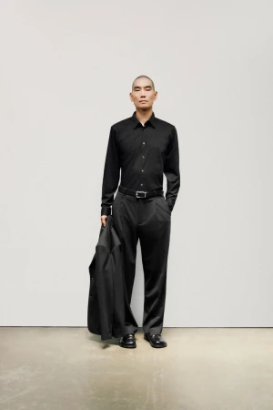 An adult Asian man with shaved head poses in an all-black satin button-down shirt, dress pants, loafers and holds a suit jacket in studio.