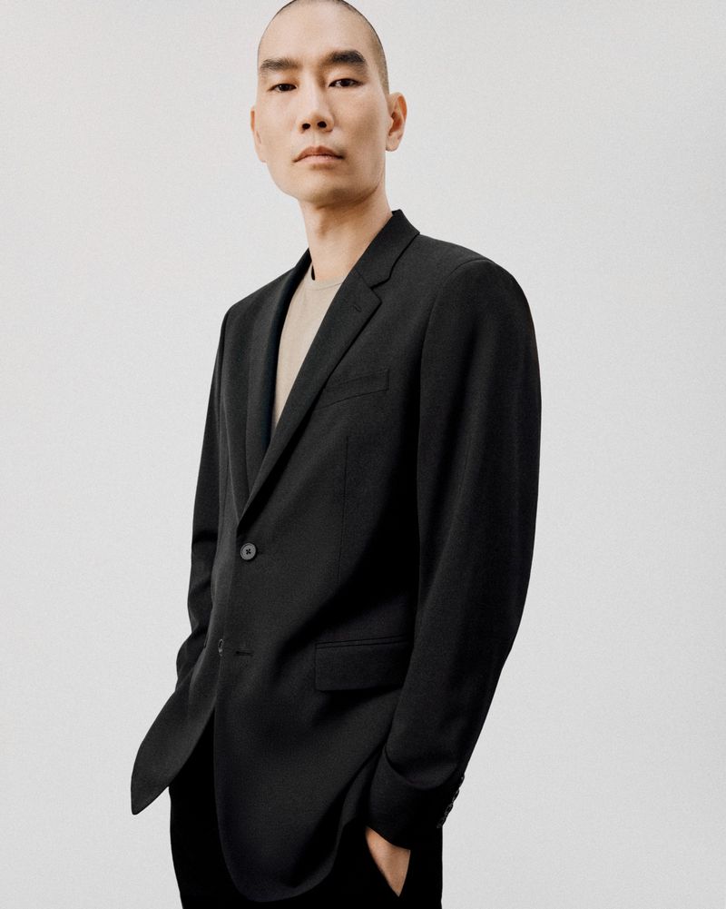 Portrait of an adult man wearing a dark gray suit jacket and a beige shirt, standing against a neutral background in a studio setting.