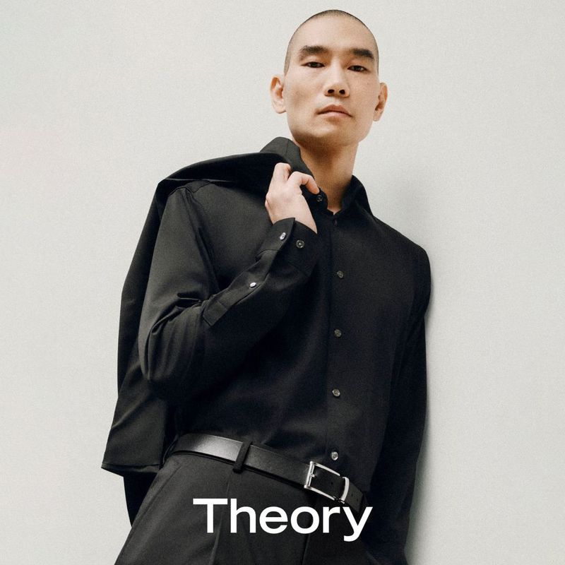 A stylish Asian male poses in a black button-down shirt, black pants, and a black belt, with a jacket draped over his shoulder.