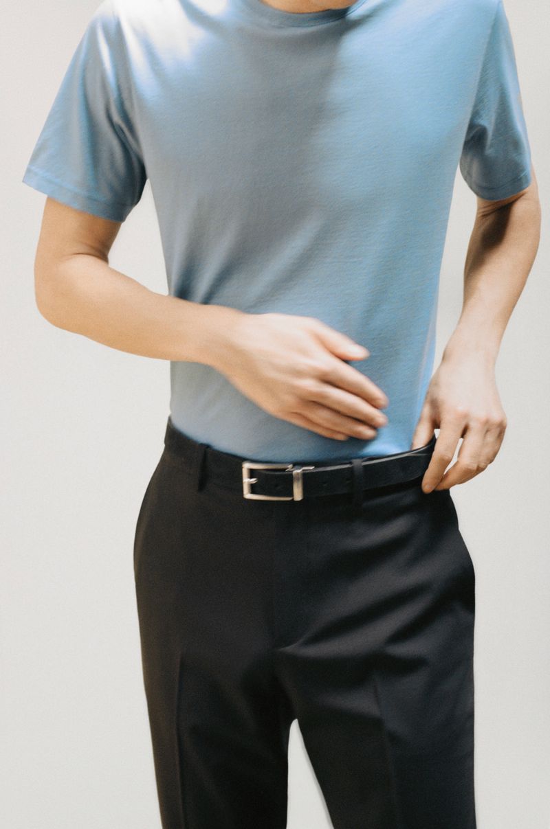 A person stands indoors wearing a blue short-sleeve shirt and black trousers with a belt, hands adjusting the belt. Neutral background.