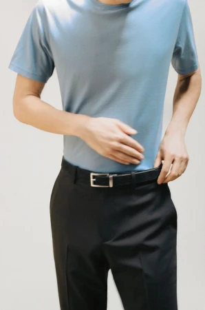A person stands indoors wearing a blue short-sleeve shirt and black trousers with a belt, hands adjusting the belt. Neutral background.