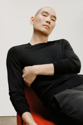 Close-up portrait of a bald Asian man wearing a black long-sleeve sweater seated against a plain white studio background.