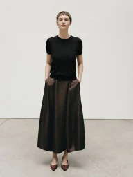 Model with short hair wearing a black t-shirt and dark midi skirt standing against a plain white studio background.