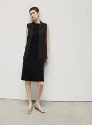 Model with short dark hair wearing a black dress and vest against a neutral background