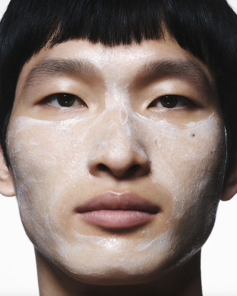 Close-up studio shot of a young adult with skincare product applied to their face, set against a clean, white backdrop.