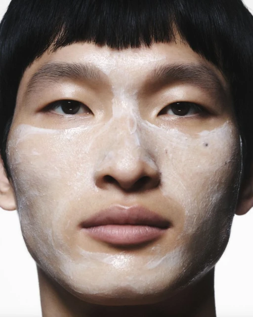 Close-up studio shot of a young adult with skincare product applied to their face, set against a clean, white backdrop.