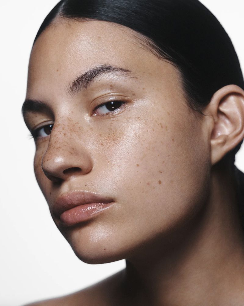 Close-up beauty shot of an adult woman with freckled skin, dark hair, light eye makeup, and bare shoulders against a plain white background.