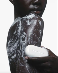 Close-up of a person with soapy suds on their shoulder holding a white bar of soap against a white background
