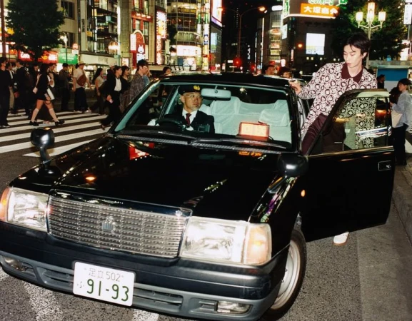 A black taxi cab awaits a passenger in a busy urban Japanese street at night with pedestrians crossing in the background.