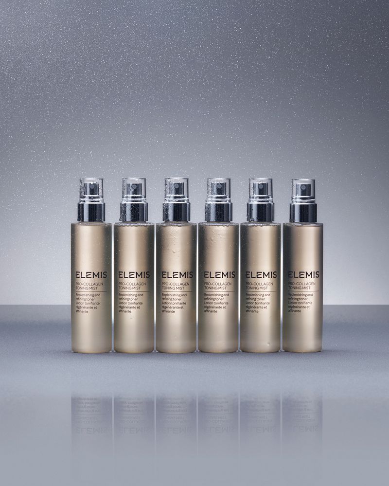 Six Elemis Pro-Collagen Toning Mist bottles in a row on a reflective surface with a neutral grey sparkling background.