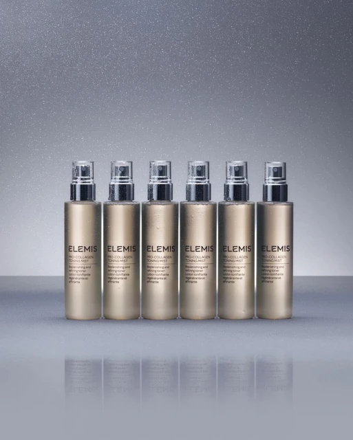 Six Elemis Pro-Collagen Toning Mist bottles in a row on a reflective surface with a neutral grey sparkling background.