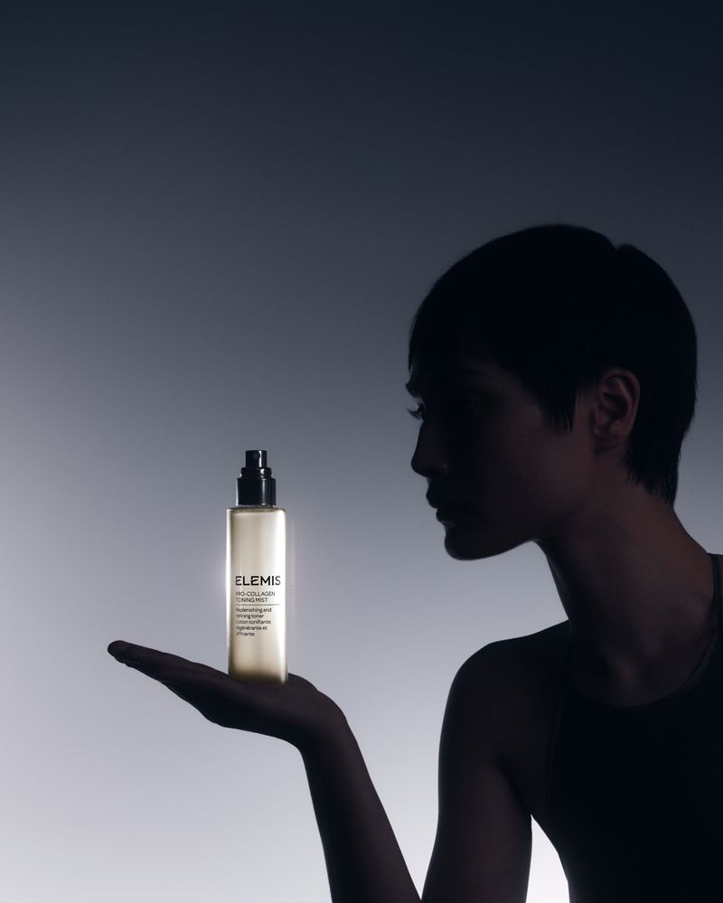 An adult female silhouette is shown from the shoulders up, holding a bottle of ELEMIS skincare product against a gradient background.