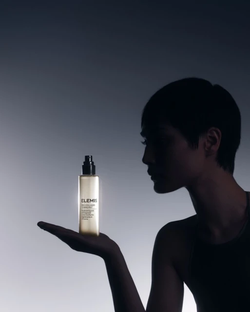 An adult female silhouette is shown from the shoulders up, holding a bottle of ELEMIS skincare product against a gradient background.