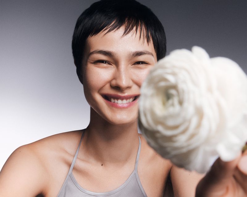 Close-up of a smiling woman with short dark hair holding a white flower against a grey gradient background in a beauty or wellness context.