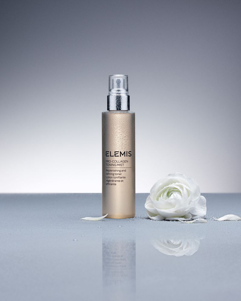 Elevated view of an Elemis Pro-Collagen Toning Mist bottle next to a white flower against a grey backdrop with reflections.