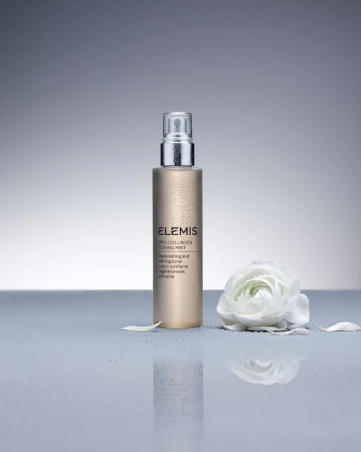 Elevated view of an Elemis Pro-Collagen Toning Mist bottle next to a white flower against a grey backdrop with reflections.