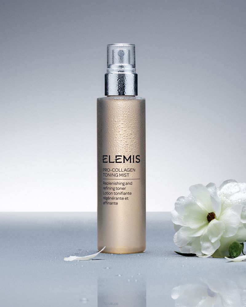 Close-up of Elemis Pro-Collagen Toning Mist spray bottle with white flower accent. Cosmetic product shot against a gray backdrop.