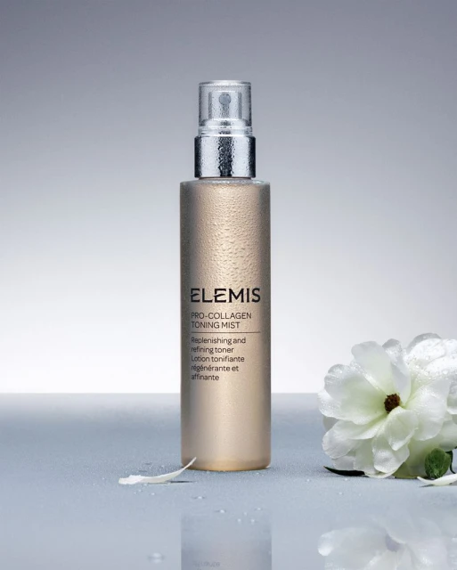 Close-up of Elemis Pro-Collagen Toning Mist spray bottle with white flower accent. Cosmetic product shot against a gray backdrop.