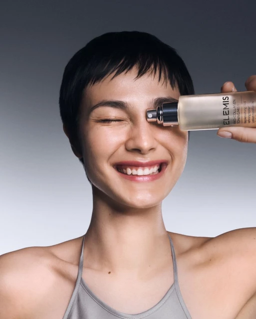 Close-up of a smiling young woman with short dark hair holding a bottle of ELEMIS skincare product to her face in a studio.