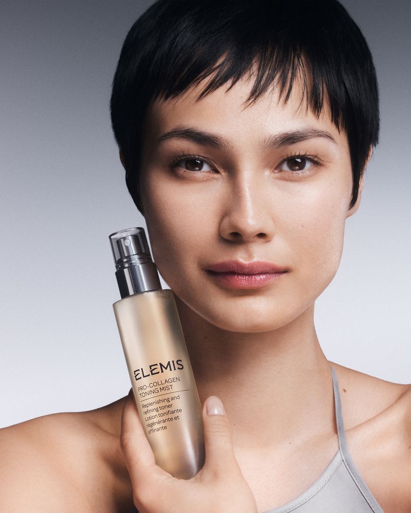 Close-up beauty shot of a woman holding Elemis Pro-Collagen Toning Mist; the product label and the model's face are clearly visible.