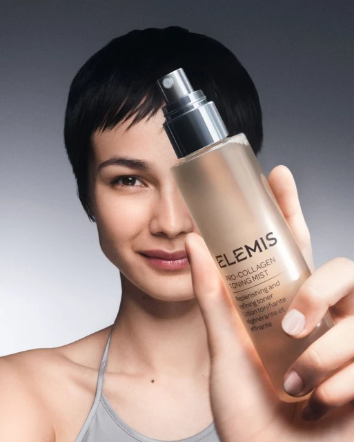 Woman poses indoors, holding a bottle of Elemis Pro-Collagen Toning Mist, wearing a light-grey tank top against a gradient-grey backdrop.