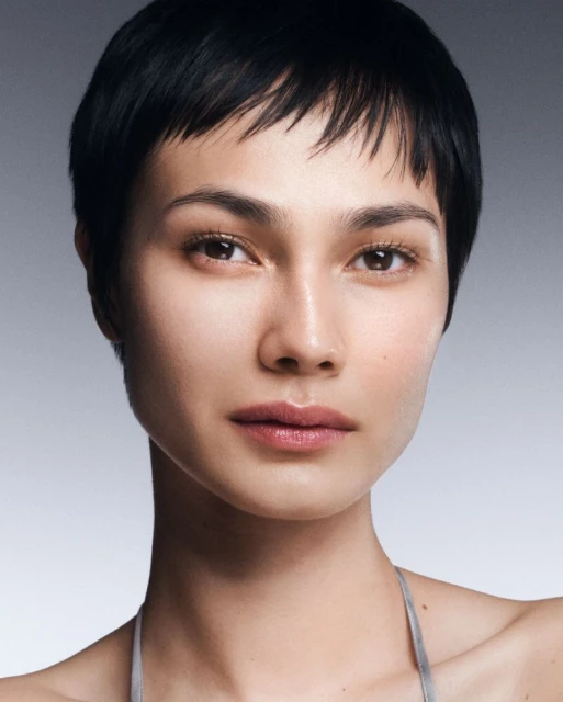 Close-up portrait of an adult woman with short, dark hair and light skin. She is wearing a silver-colored top and looking forward.