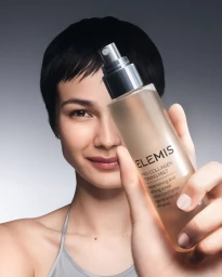 Model holding Elemis toning mist bottle close to face against a clean grey studio background