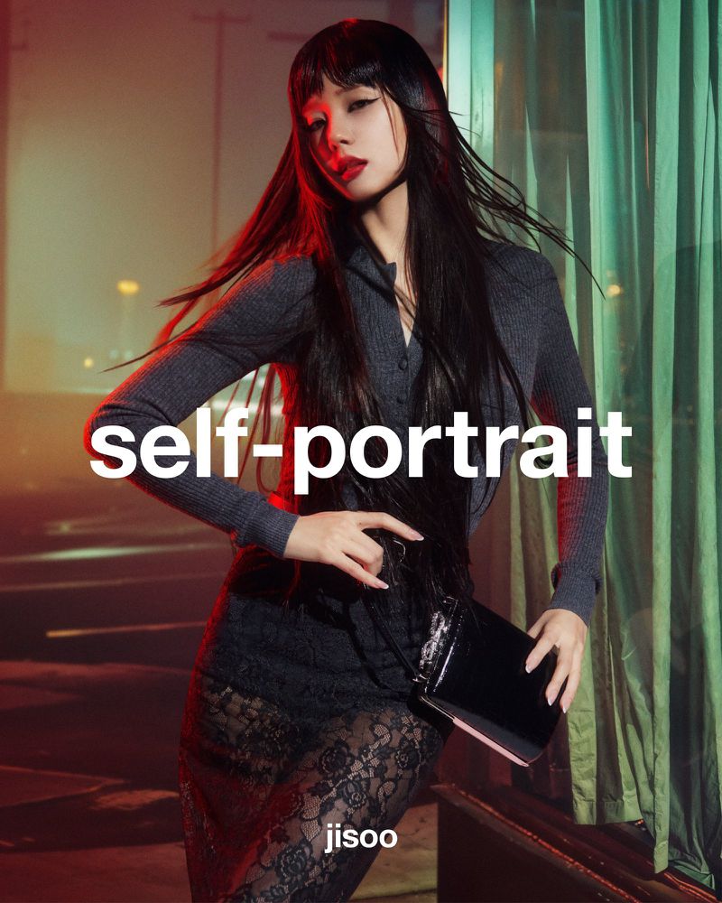 Fashion portrait of adult female model with long, dark hair, wearing grey form-fitting top and lace skirt, holding a black bag.