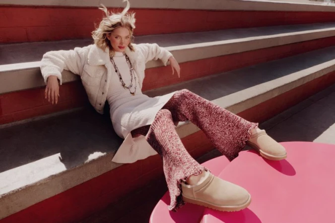 A blonde model poses on red and white steps wearing a white jacket, dress, mauve pants, and beige UGG boots for a fashion shoot.