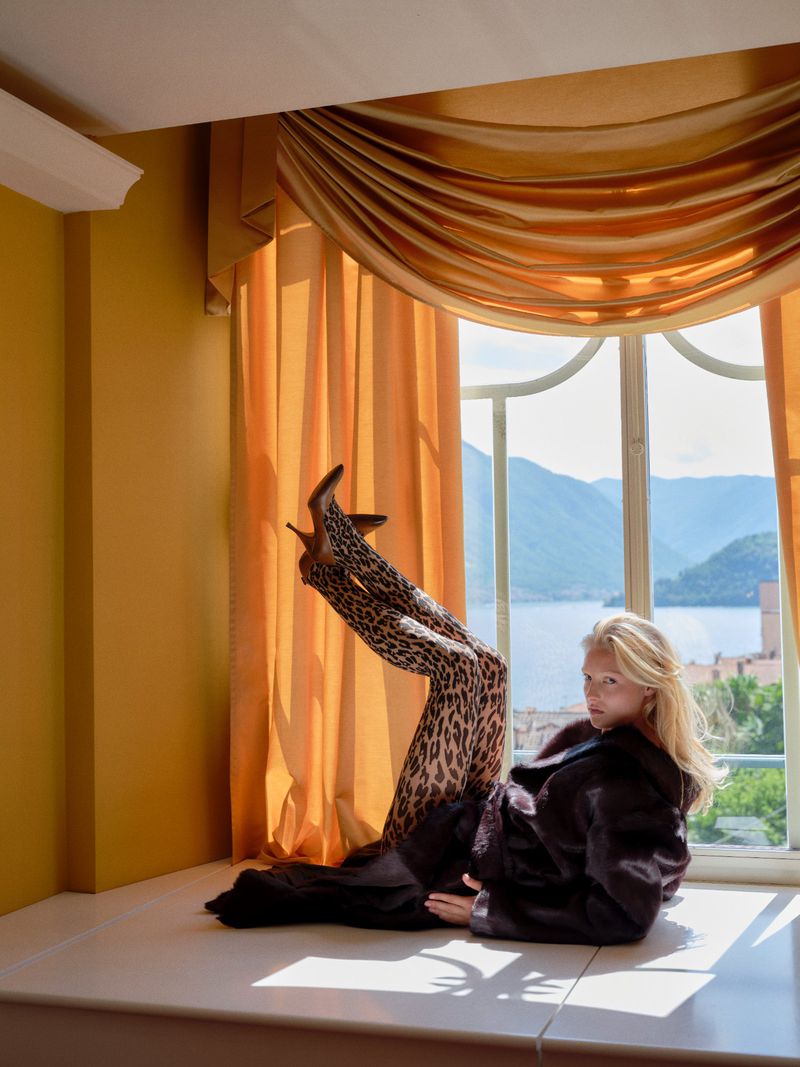 Fashionable woman with blonde hair wearing animal print leggings and high heels, posing in front of a bright window with a lake view.