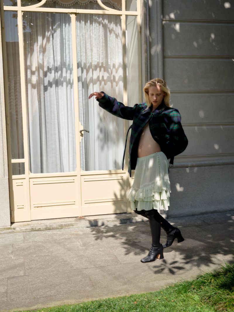 A slender blonde woman poses in front of a building with a window, wearing a short skirt, boots, and a plaid blazer in natural light.