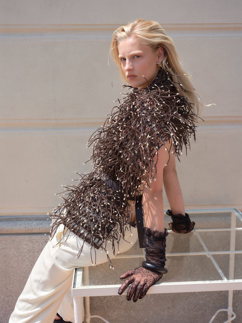 A fair-skinned woman with blonde hair in a feathered top and cream pants leans against a white grid structure in outdoor lighting.